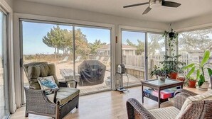 Una habitación amplia con grandes puertas corredizas de vidrio que dan a un patio trasero, ventilador de techo, mobiliario de mimbre y una mesita con libros.