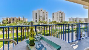 A balcony with a table, a bottle, and glasses, overlooking a street with palm trees and buildings.
