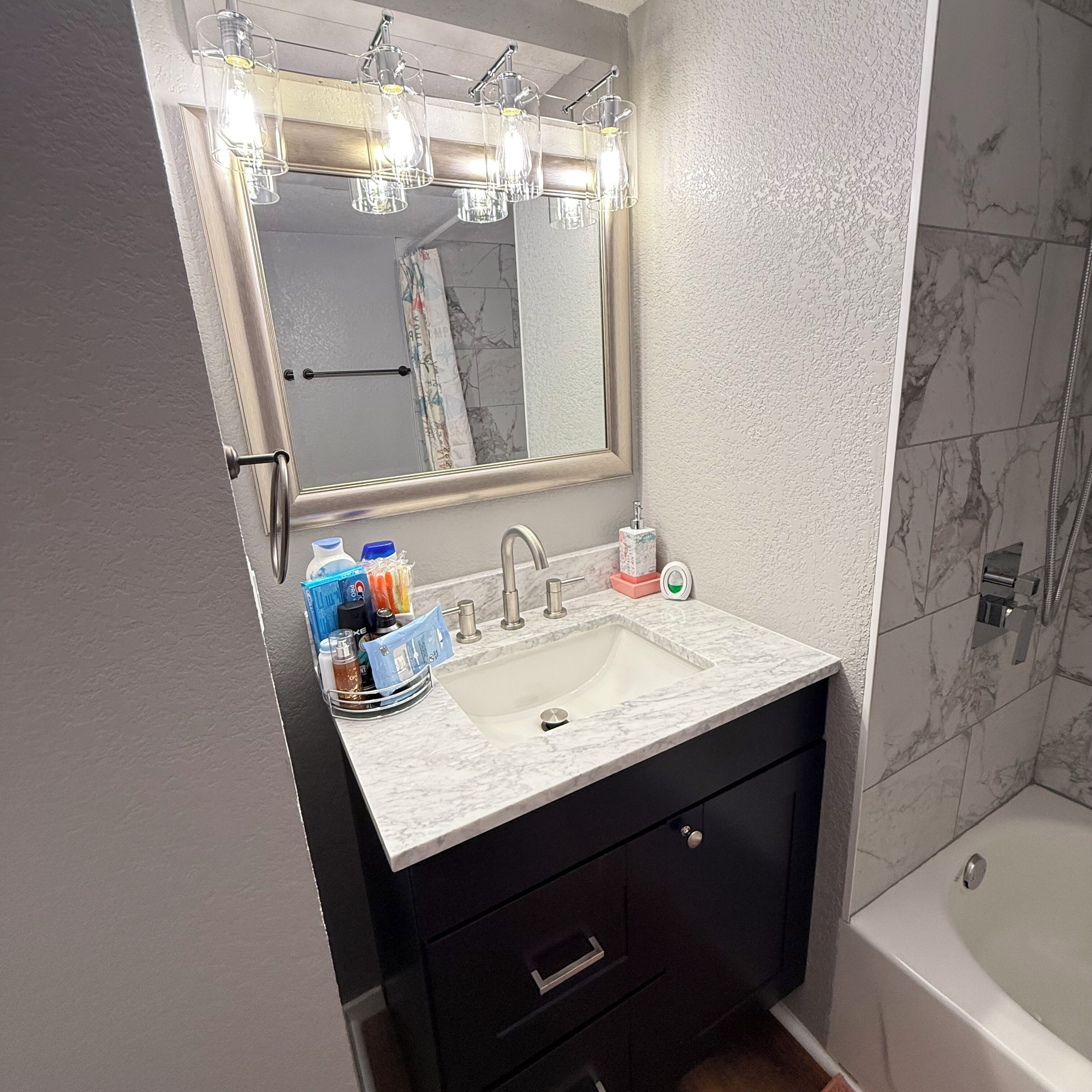 A bathroom with a marble sink, a mirror with lights, a black vanity, and a shower area.