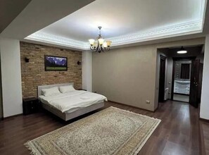 A bedroom with a bed, a chandelier, a television on the wall, and a hallway leading to a bathroom. - Lite Guest House (Bishkek)
