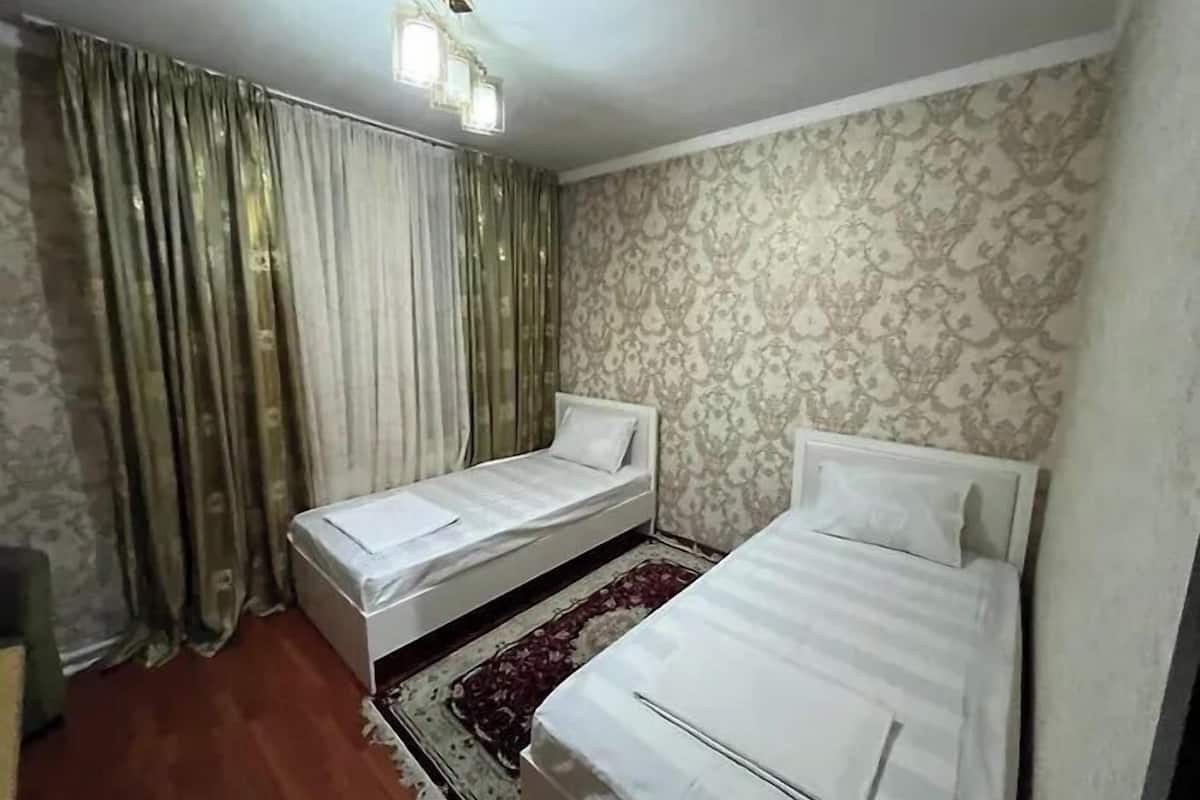 A room with two single beds, a patterned wallpaper, a patterned rug, and a ceiling light fixture.