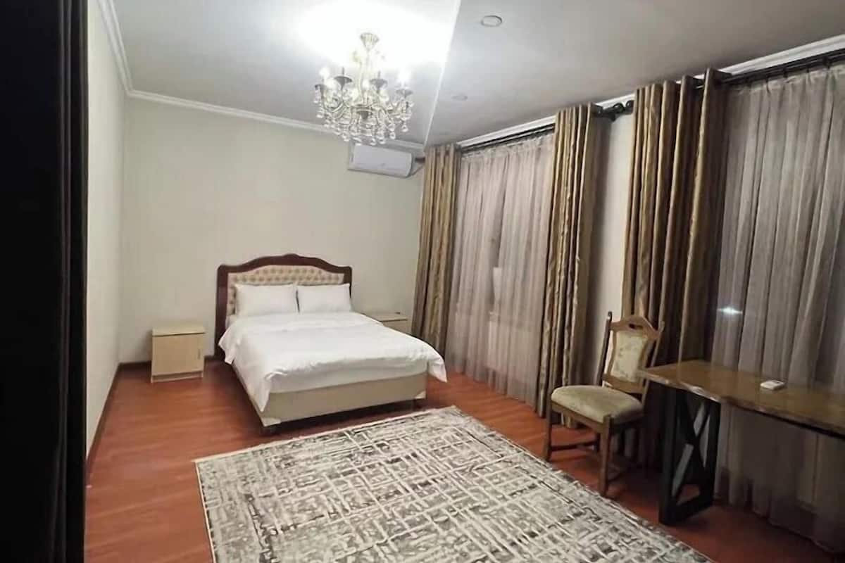 A hotel room with a bed, a desk, a chair, a chandelier, and a large window with curtains.