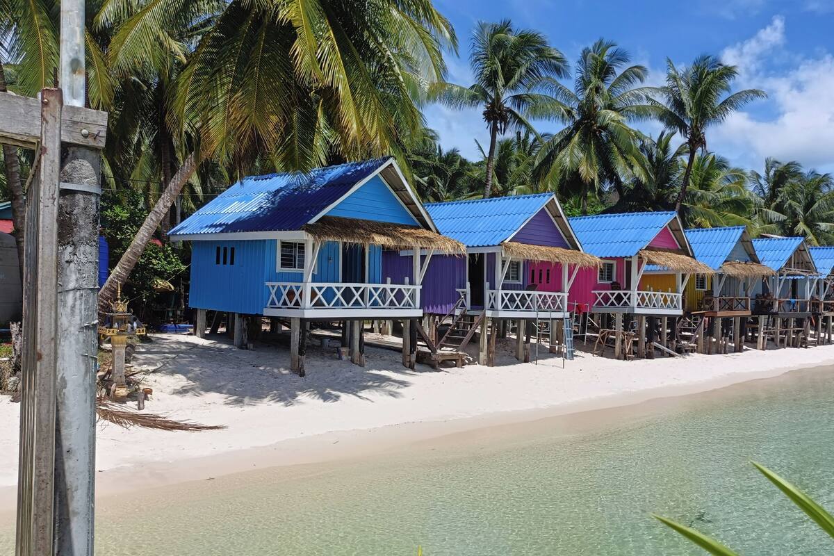 A row of colorful beach huts with thatched roofs, wooden stilts, and white railings, situated on a sandy beach with clear turquoise water and palm trees in the background.