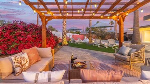 Een buitenpatio met een houten pergola, lichtslingers en een vuurkorf.