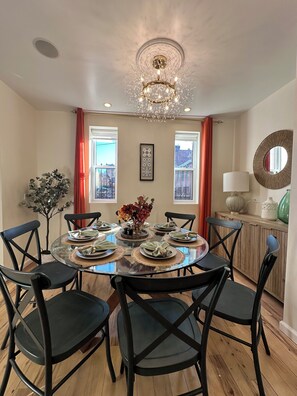 A dining room with a round glass table, black chairs, a chandelier, and a view through large windows with red curtains.