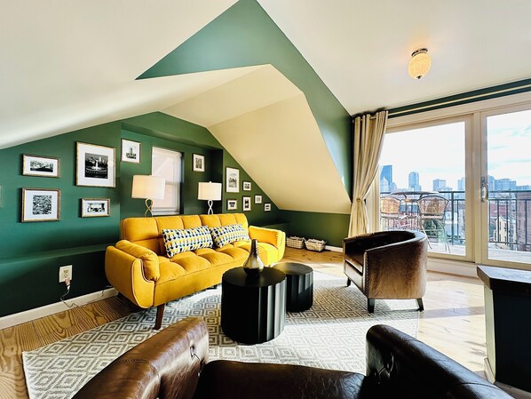 A modern living room with a yellow sofa, black coffee table, and leather armchairs. There are framed pictures on the green walls and a city view through the large window.