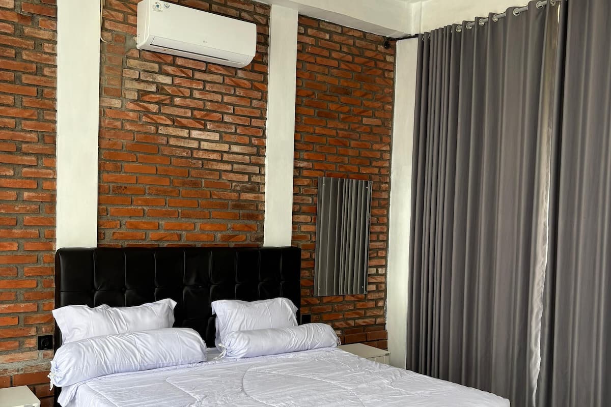 Un lit bien fait, avec du linge de lit blanc, une tête de lit noire et un mur en briques sur lequel est installé un appareil de climatisation.