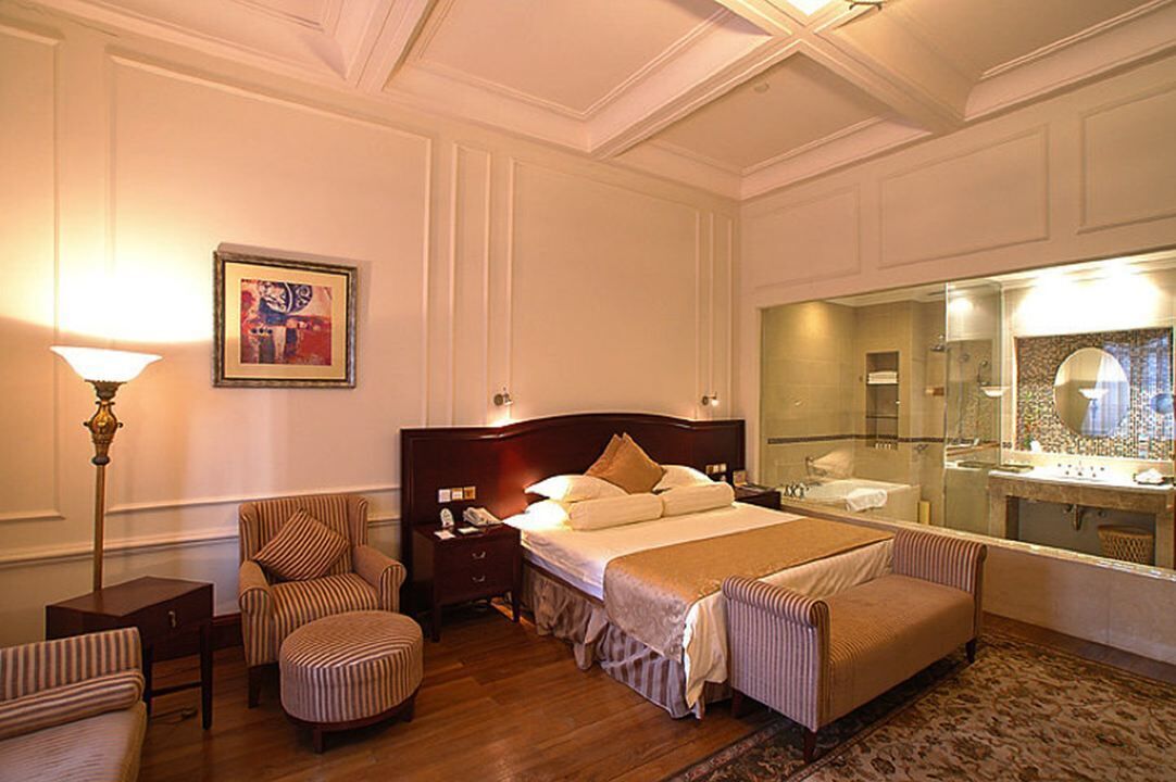 Une chambre d’hôtel dotée d’un grand lit, d’un coin salon avec un fauteuil et un pouf, d’une table de chevet avec une lampe et d’un grand miroir au-dessus du lavabo.
