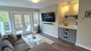 Una sala de estar moderna con un sofá seccional, un televisor de pantalla plana y una zona de cocina con gabinetes grises.