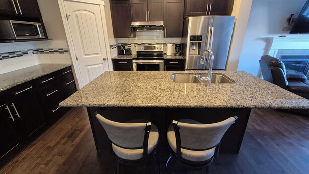 A modern kitchen with dark wood cabinets, stainless steel appliances, granite countertops, and a central island with bar stools. - Sean Comfy Home Modern Escape in Edmonton – Near Northgate  (Edmonton)