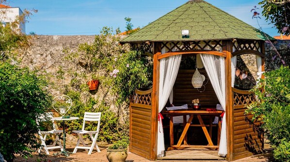 A wooden gazebo with a tiled roof, a table with chairs, and a potted plant.