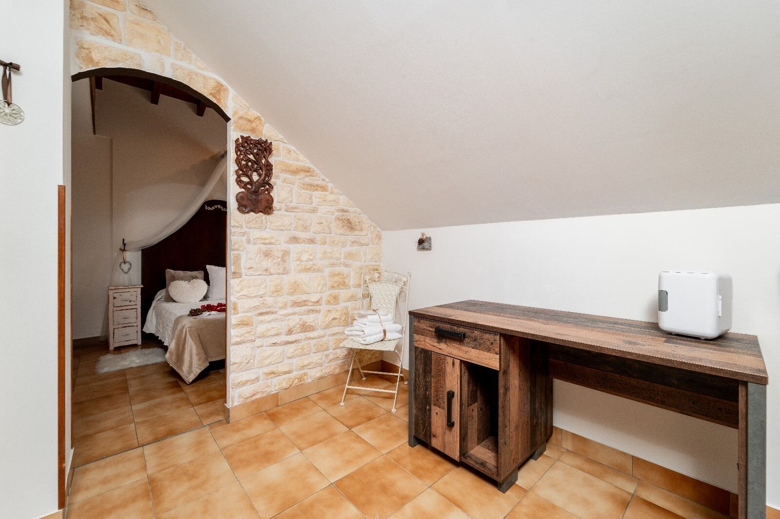 A bedroom with a stone wall, a wooden desk, and a white bedside table.
