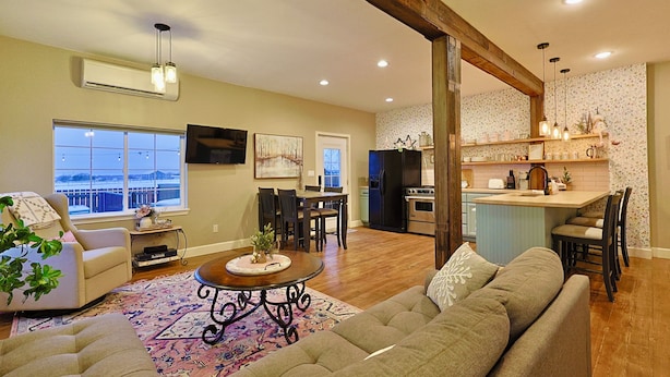 A spacious living room with a sofa, a coffee table, and a dining area with chairs. There is a kitchen area with a refrigerator and a sink.