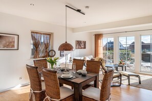 A dining area with a table set for six, surrounded by wicker chairs, a hanging light fixture, and large windows with curtains. - Holiday Home 'Casa Meeresbrise am Vogelpark Marlow' with Terrace, Hot Tub & Garden (Marlow)