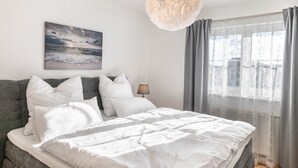 Ein Schlafzimmer mit einem Bett, Kissen, einer Lampe, einem Gemälde und einem Fenster mit Vorhängen.
