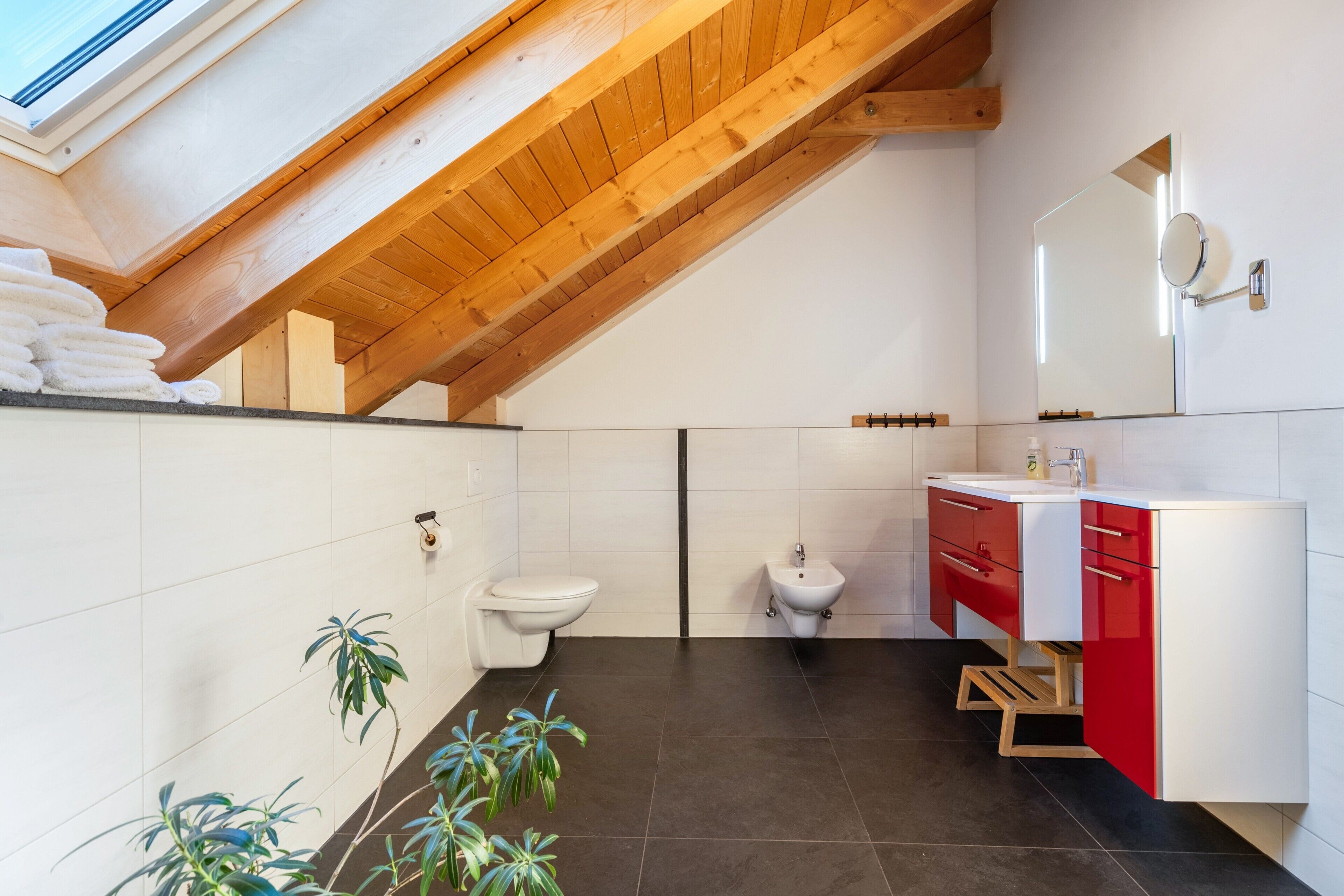 Een moderne badkamer met dakraam, witte tegels, een wastafel in rood en wit, een douchetoilet en twee toiletten.