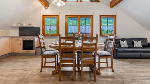 A dining area with a wooden table and chairs, a kitchen with a stove, and a black leather sofa. - Holiday Apartment 'Sum' with Mountain View, Shared Garden, and Wi-Fi (Oberwolfach)