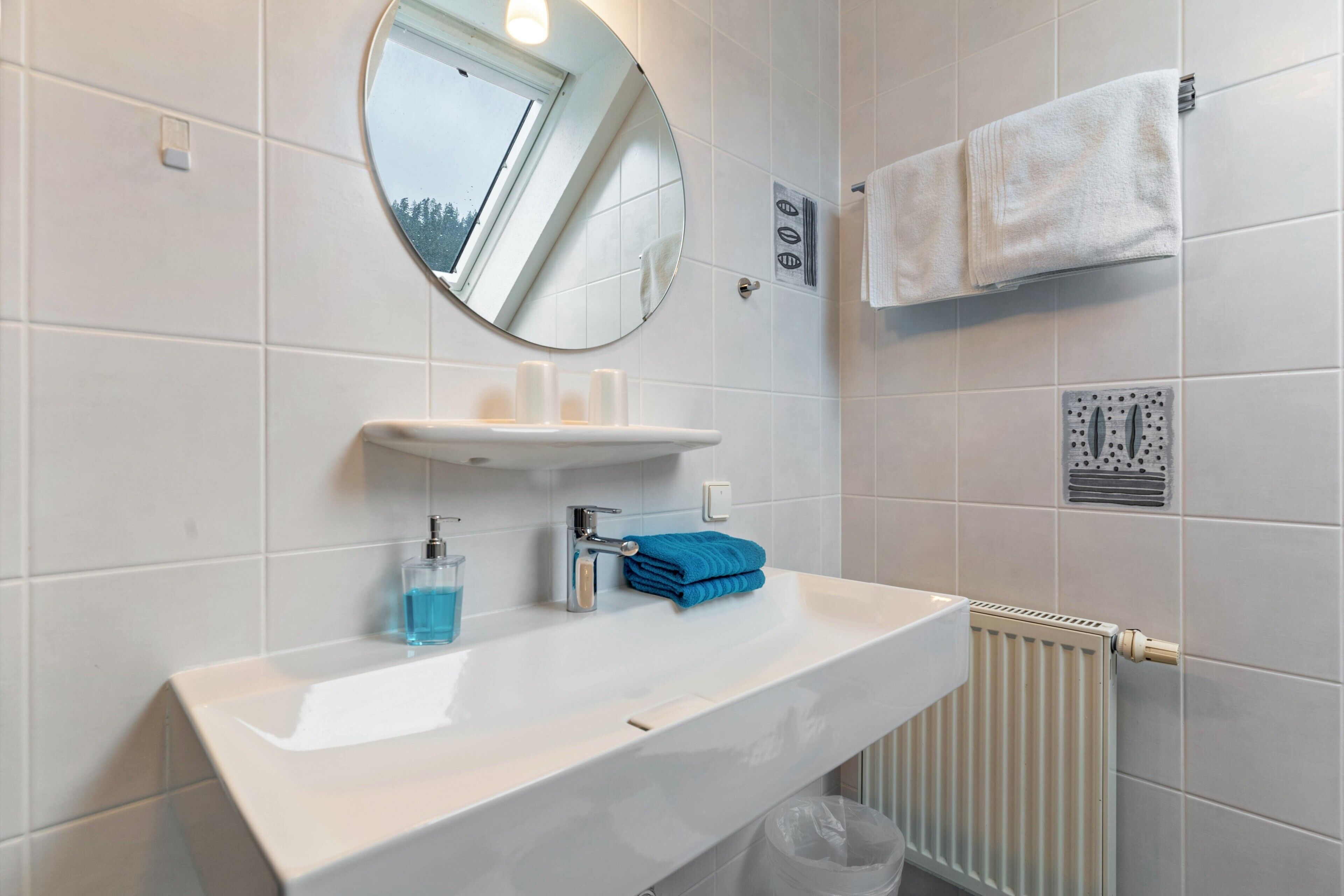 Un baño con un lavamanos blanco, un espejo, un perchero para toallas y una ventana.