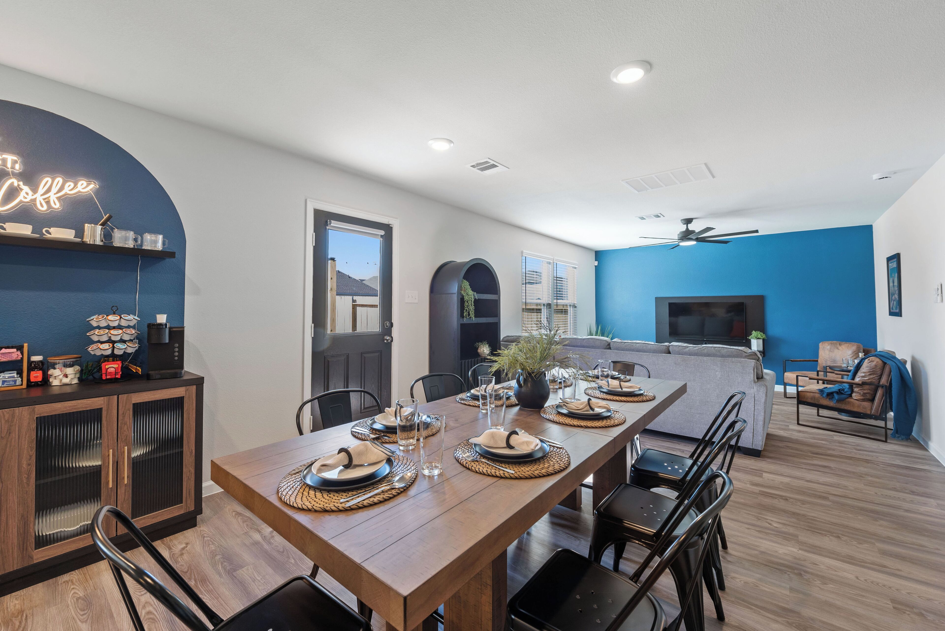 A modern dining area with a wooden table set for four, a neon sign reading "Coffee," and a kitchen in the background.