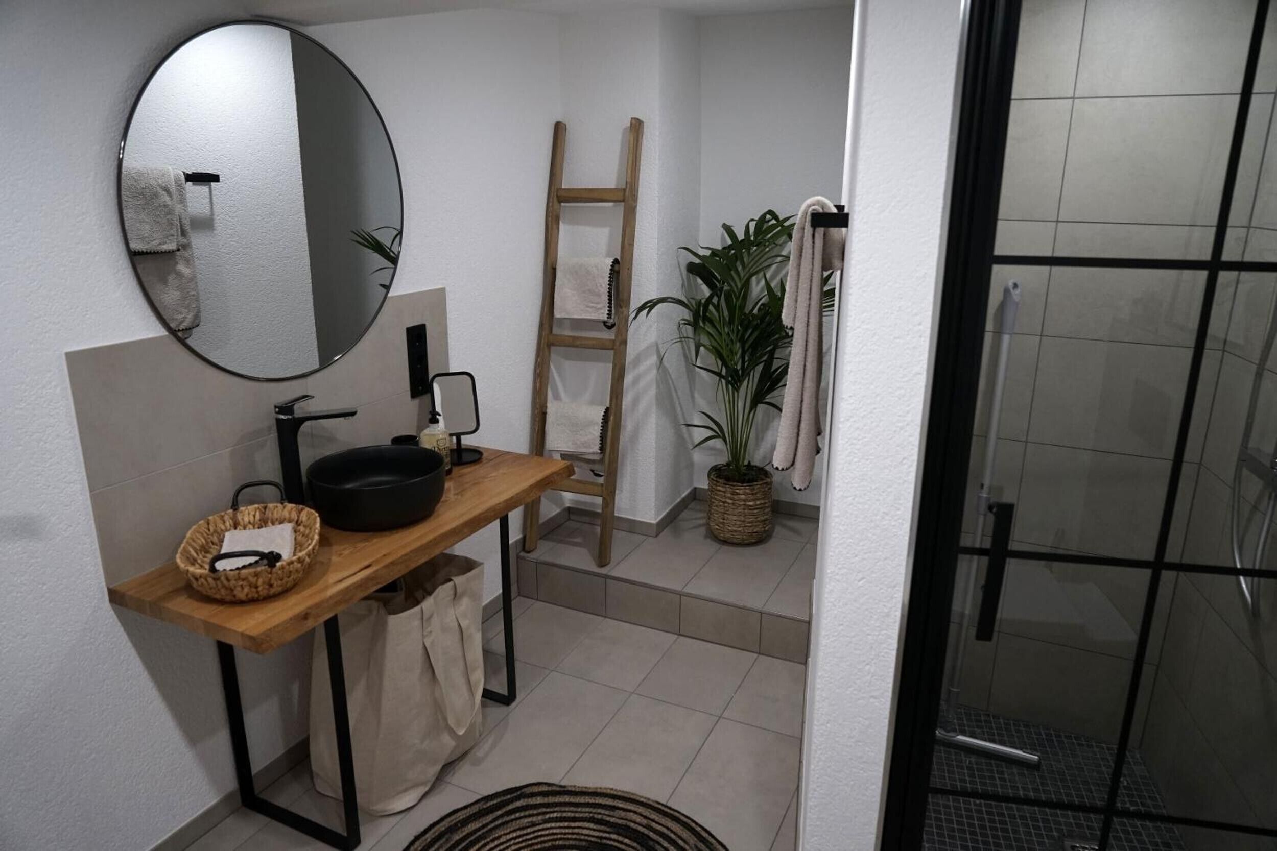 Un bagno con un mobile lavabo in legno, uno specchio rotondo, un porta asciugamani, una scala, una pianta in vaso e uno spazio doccia.