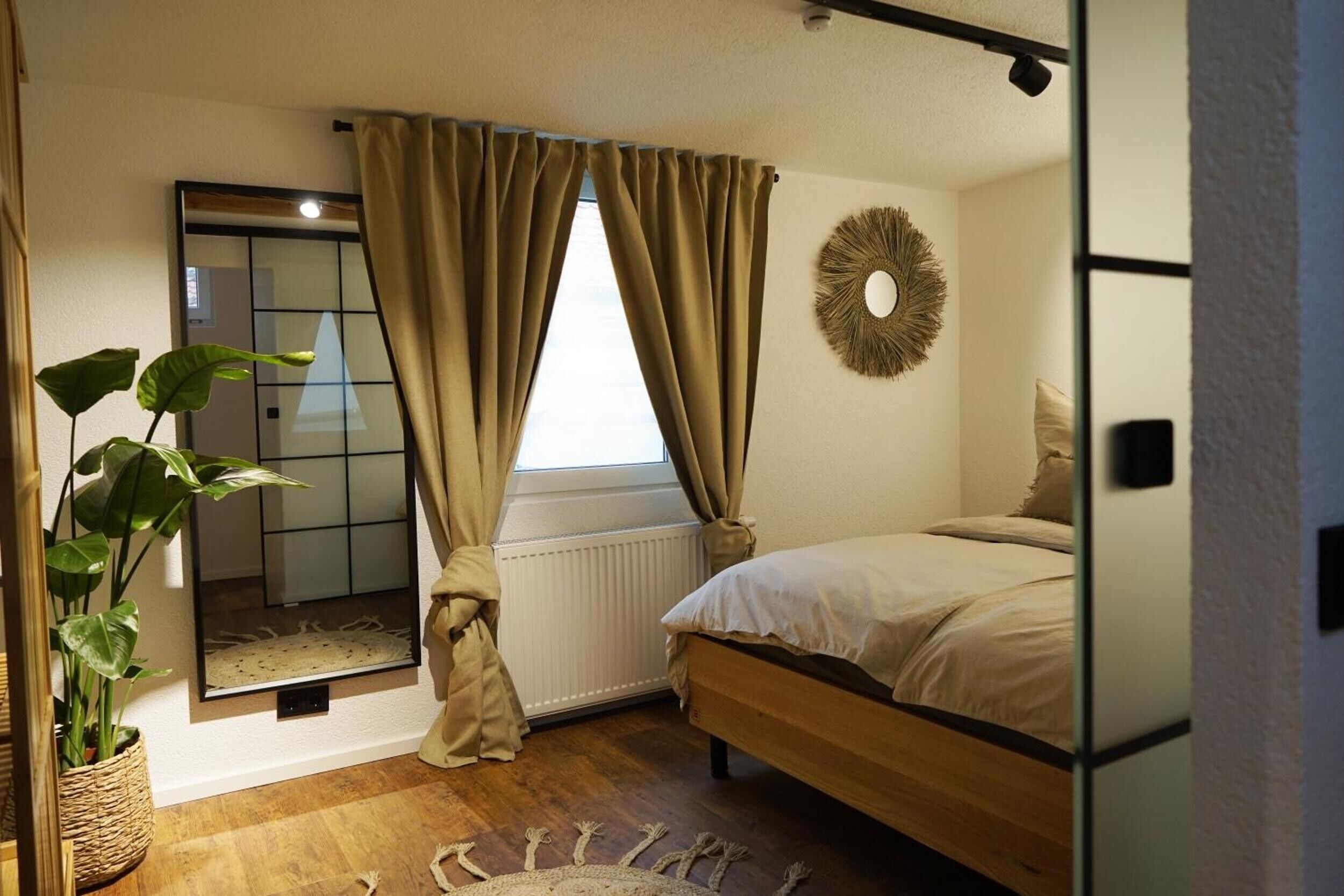 A bedroom with a bed, a mirror, a radiator, a window with curtains, and a plant.