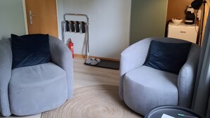 Een kamer met twee grijze fauteuils, een rond vloerkleed, een kleine tafel met een papier, een kapstok en een deur.