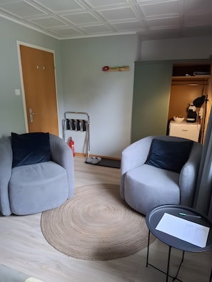 Una habitación con dos sillones grises, un tapete redondo, una mesita con un papel, un perchero y una puerta.