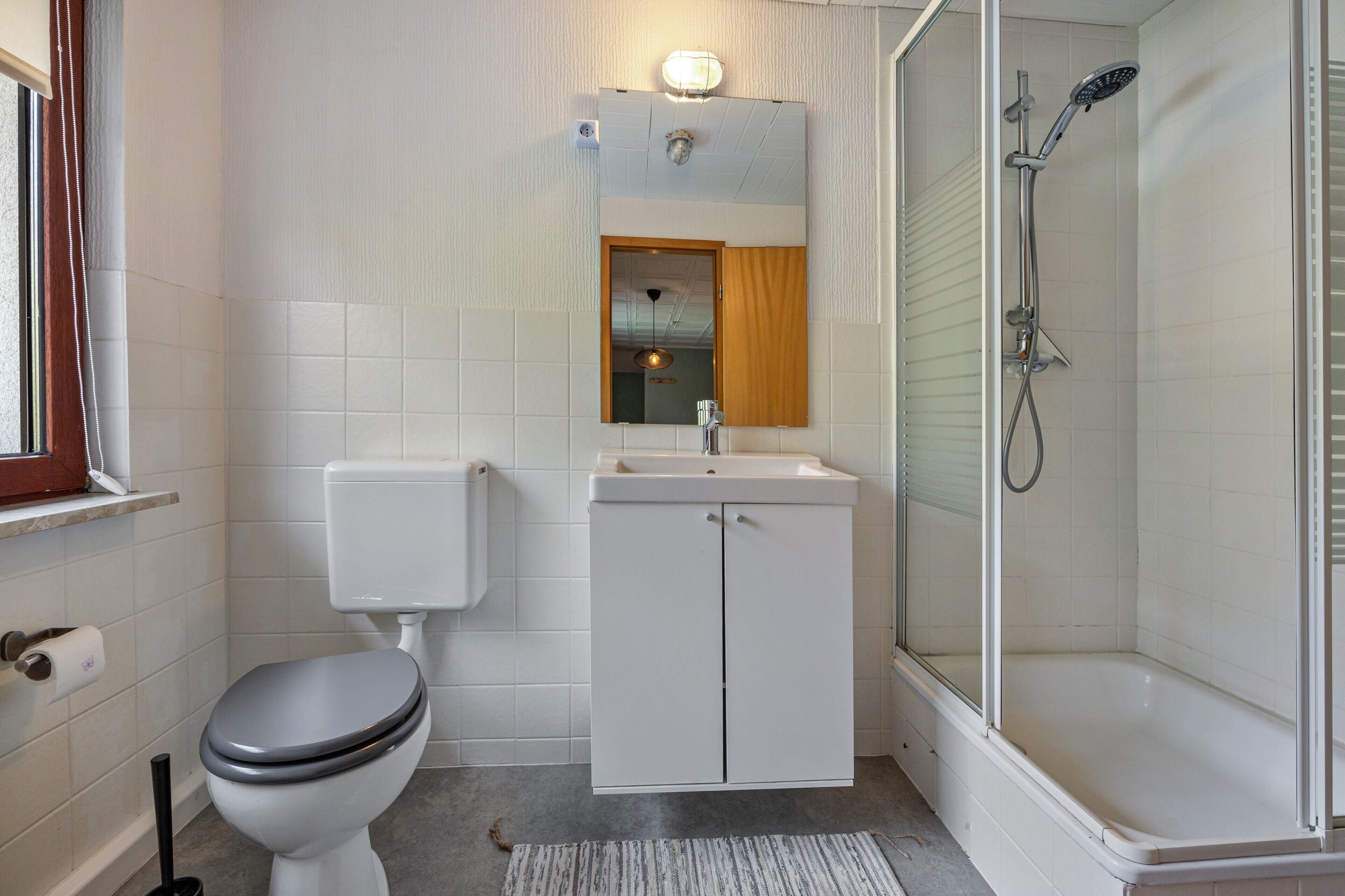 A bathroom with a toilet, sink, mirror, shower, and a window.