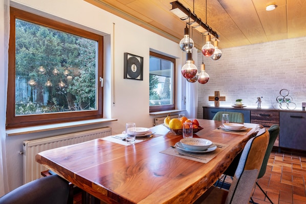 Un comedor con una mesa de madera, sillas y un cuenco de fruta. Se ven ventanas con vistas a árboles y un muro de ladrillo.