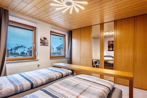 Un dormitorio con dos camas, un armario de madera y una ventana con vistas a casas.