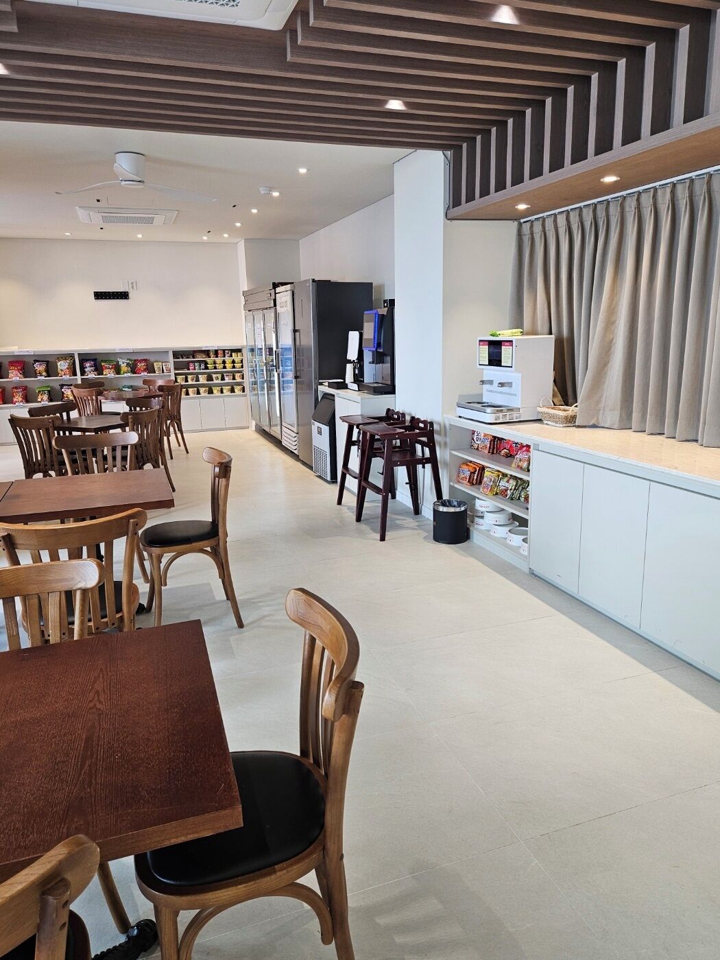 A modern cafe with wooden tables and chairs, a counter with a coffee machine, and refrigerated display cases in the background.