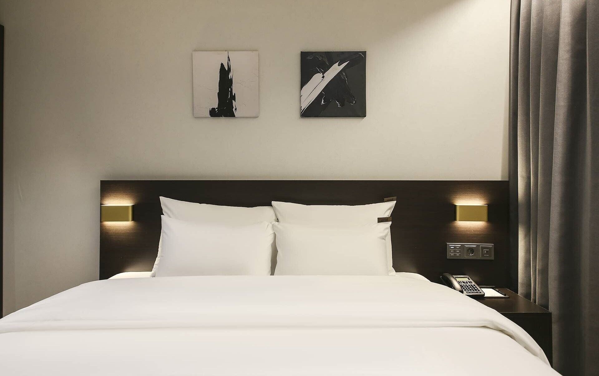 Een hotelkamer met een groot bed, twee bedlampen, een nachtkastje met een telefoon en twee aan de muur bevestigde kunstwerken.