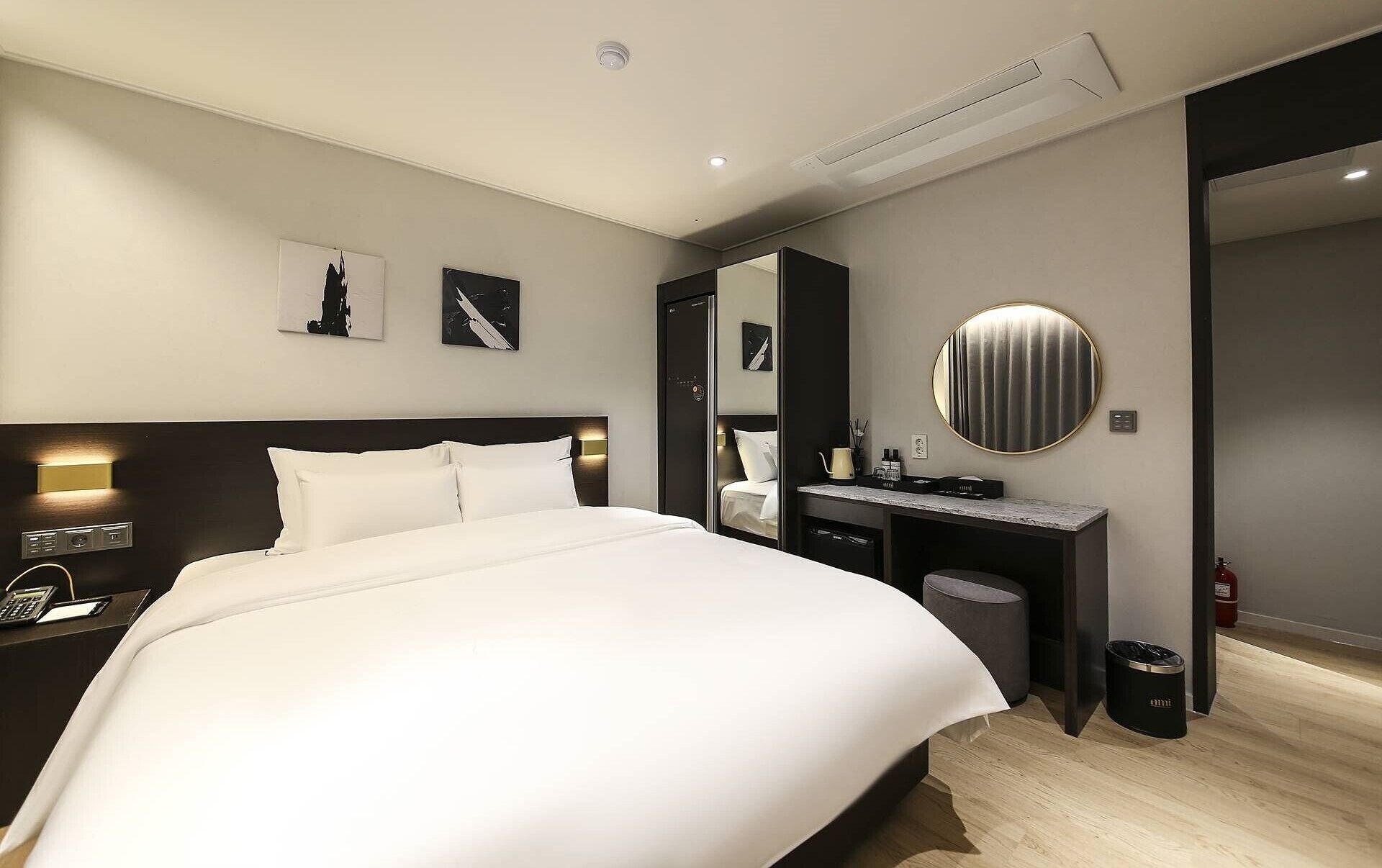 A modern hotel room with a large bed, a desk with a computer, a mirror, and a round wall clock.