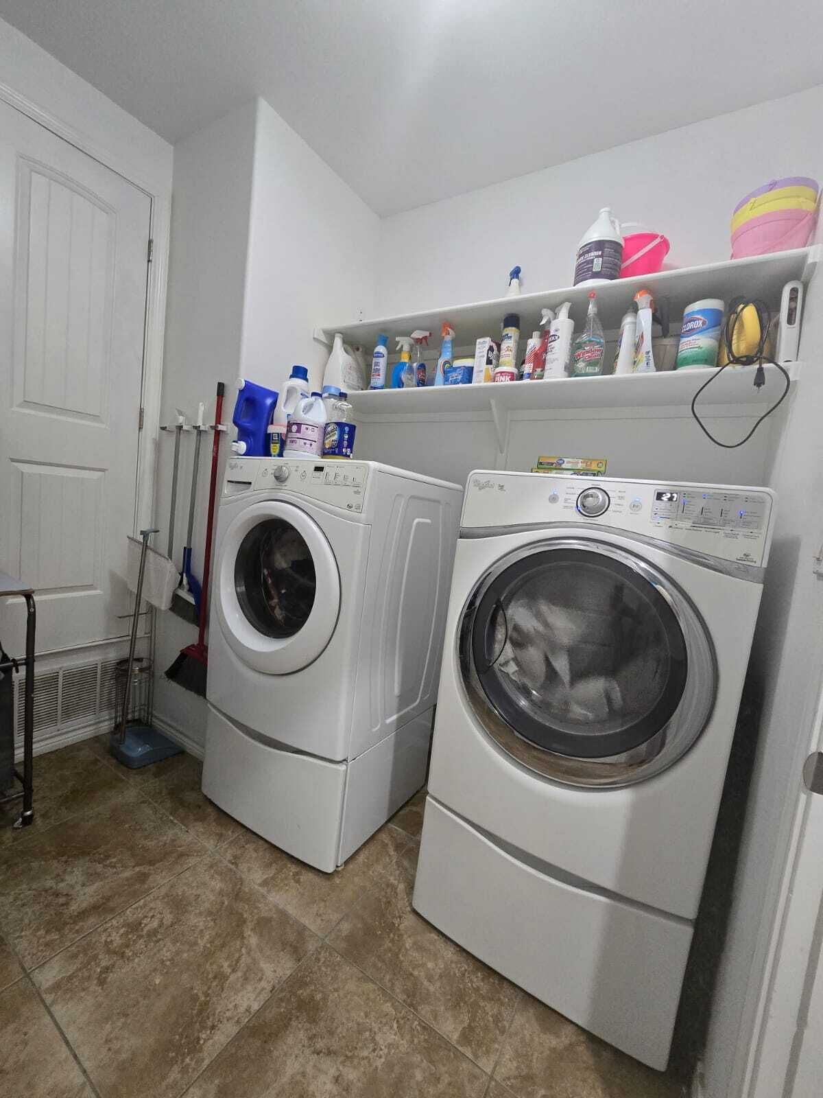 A laundry room with two front-loading washing machines, a shelf with cleaning supplies, and a mop and broom.