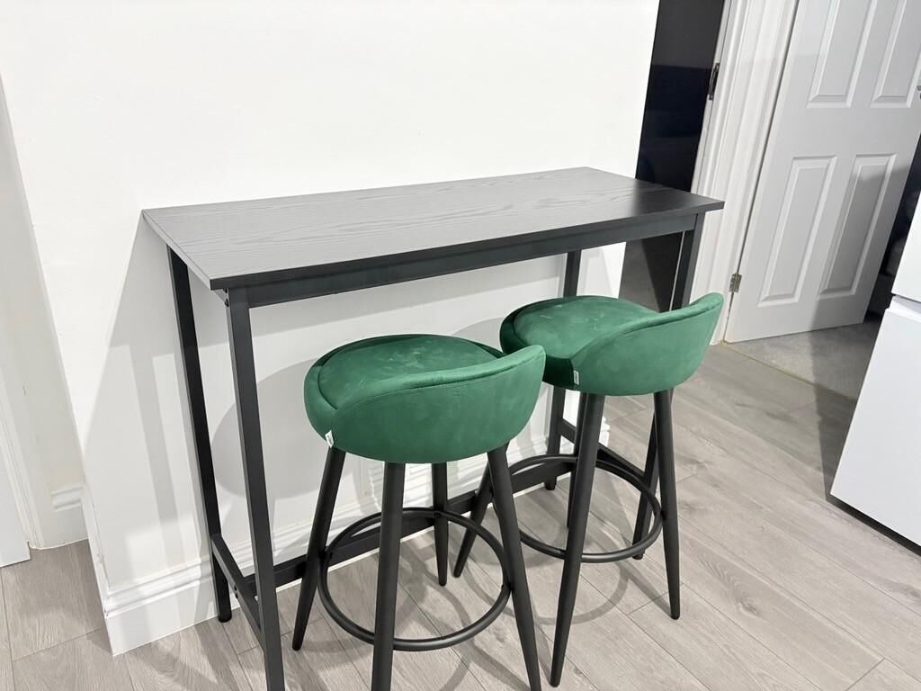 Et lille tekøkken med et mørkt bord og to grønne barstole.