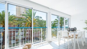 Un balcon lumineux et spacieux, doté de grandes fenêtres, de meubles blancs et offrant une vue sur les palmiers et les immeubles.