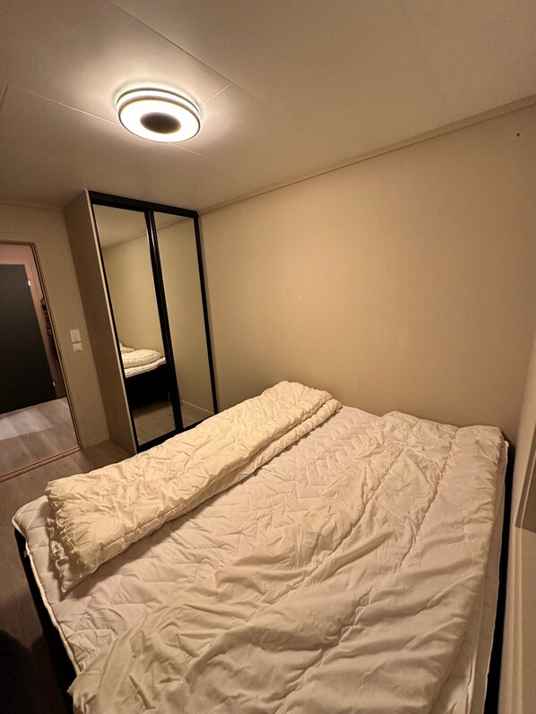 A small, single-bed room with a white bedspread, a mirror, and a ceiling light.