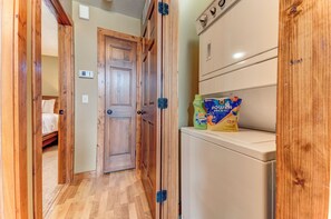 Een smalle gang met houten deuren die leiden naar een slaapkamer met een bed. Een wasmachine en wasmiddel staan op een plank.