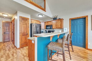 A kitchen with wooden cabinets, a stainless steel refrigerator, a microwave, and a bar area with woven chairs.