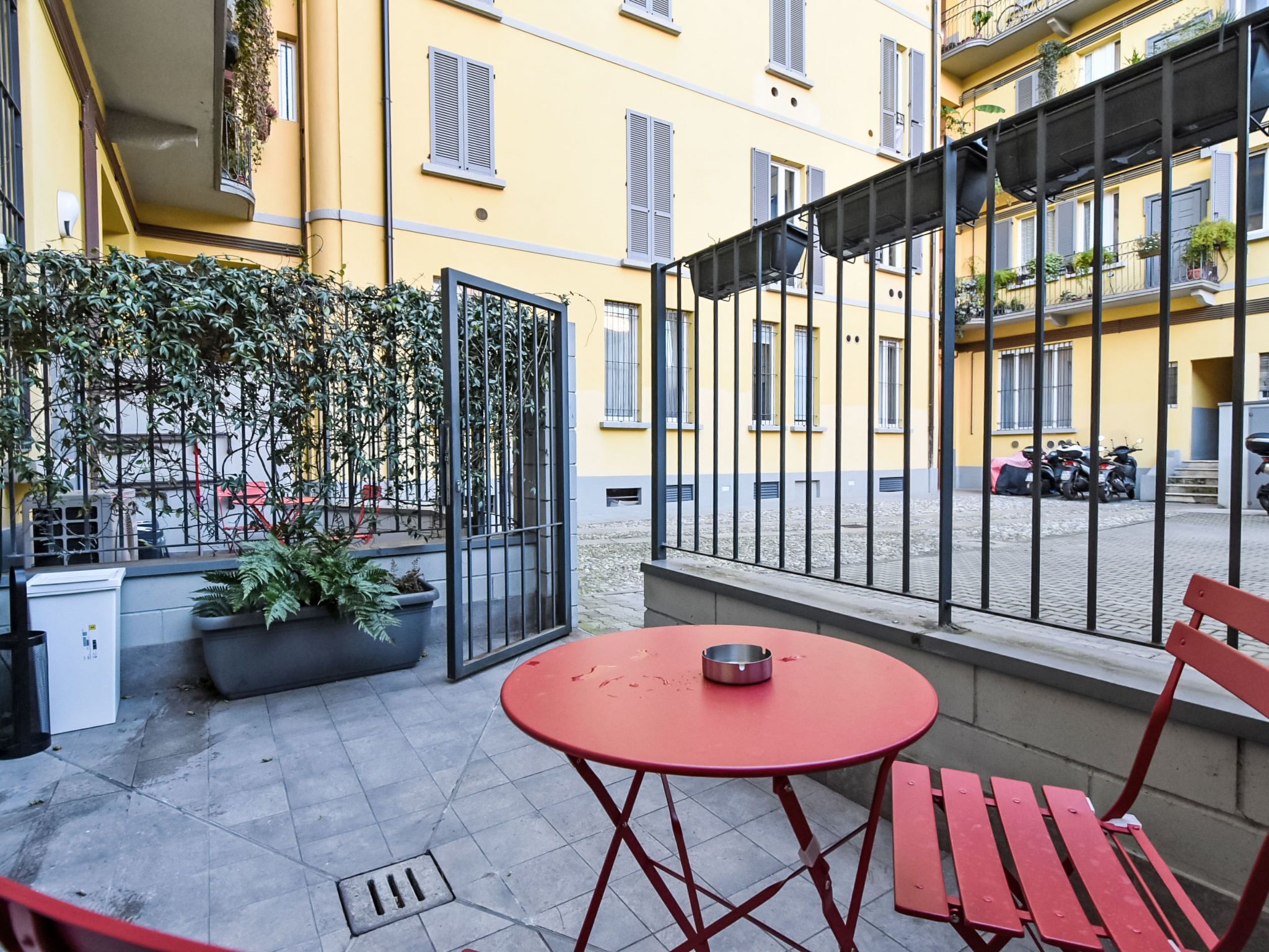 A courtyard with a red table and bench, potted plants, and a metal fence.