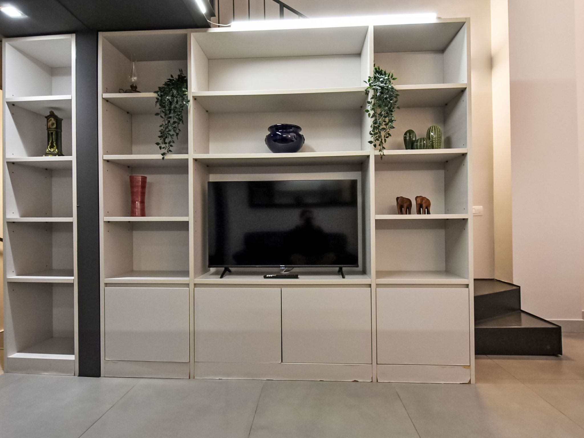 A modern living room with a flat-screen TV on a white entertainment unit, surrounded by shelves with various decorative items.