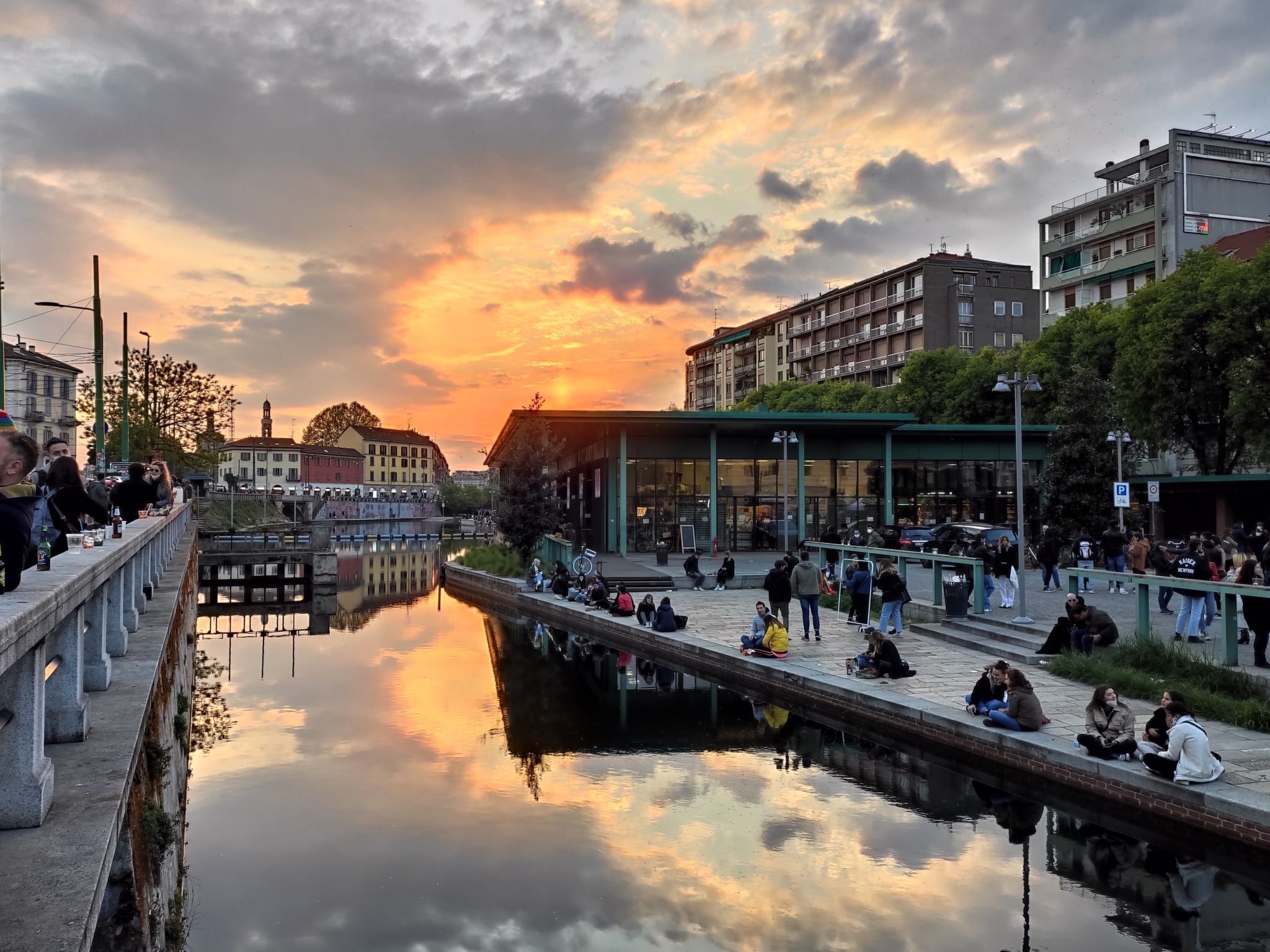 Un paseo ribereño con gente sentada y caminando, rodeado de edificios y un cielo al atardecer.