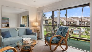 A modern living room with a sofa, a glass coffee table, a wicker chair, and a view of a pool and palm trees through large windows.