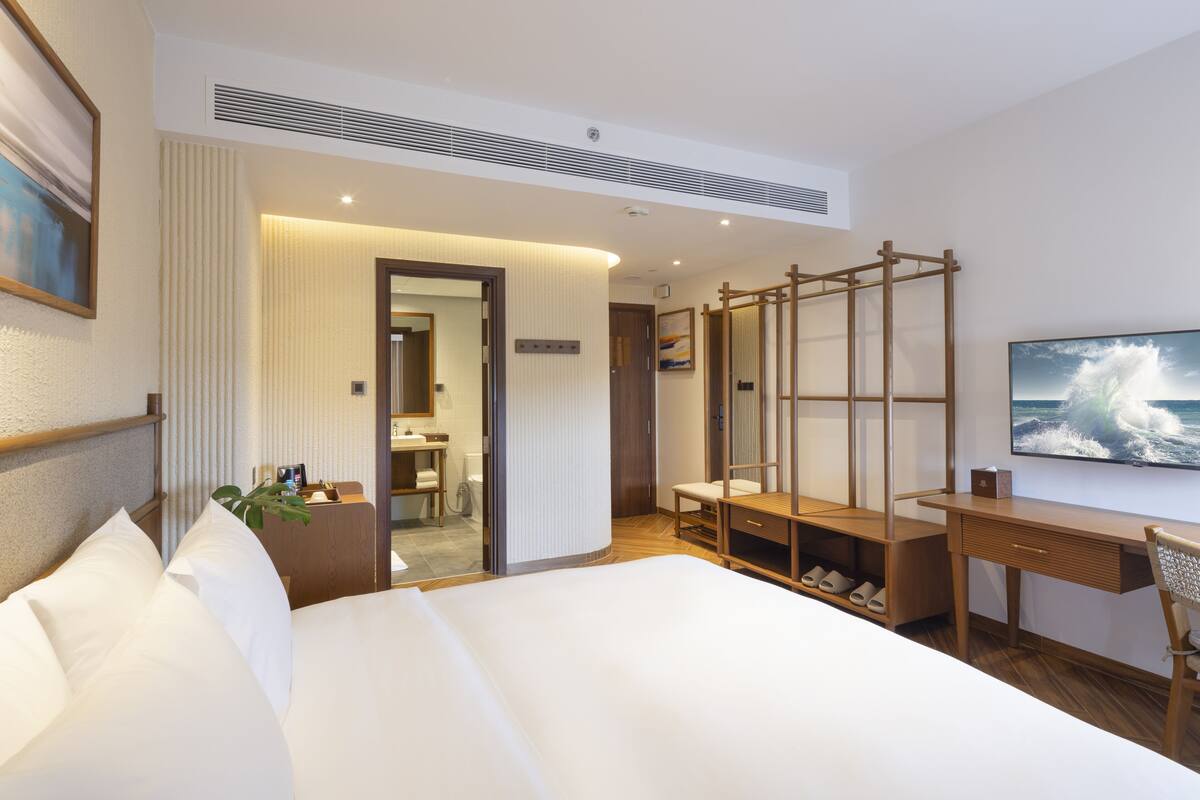 A hotel room with a large bed, a desk with a chair, a TV, and a bathroom visible through an open door.