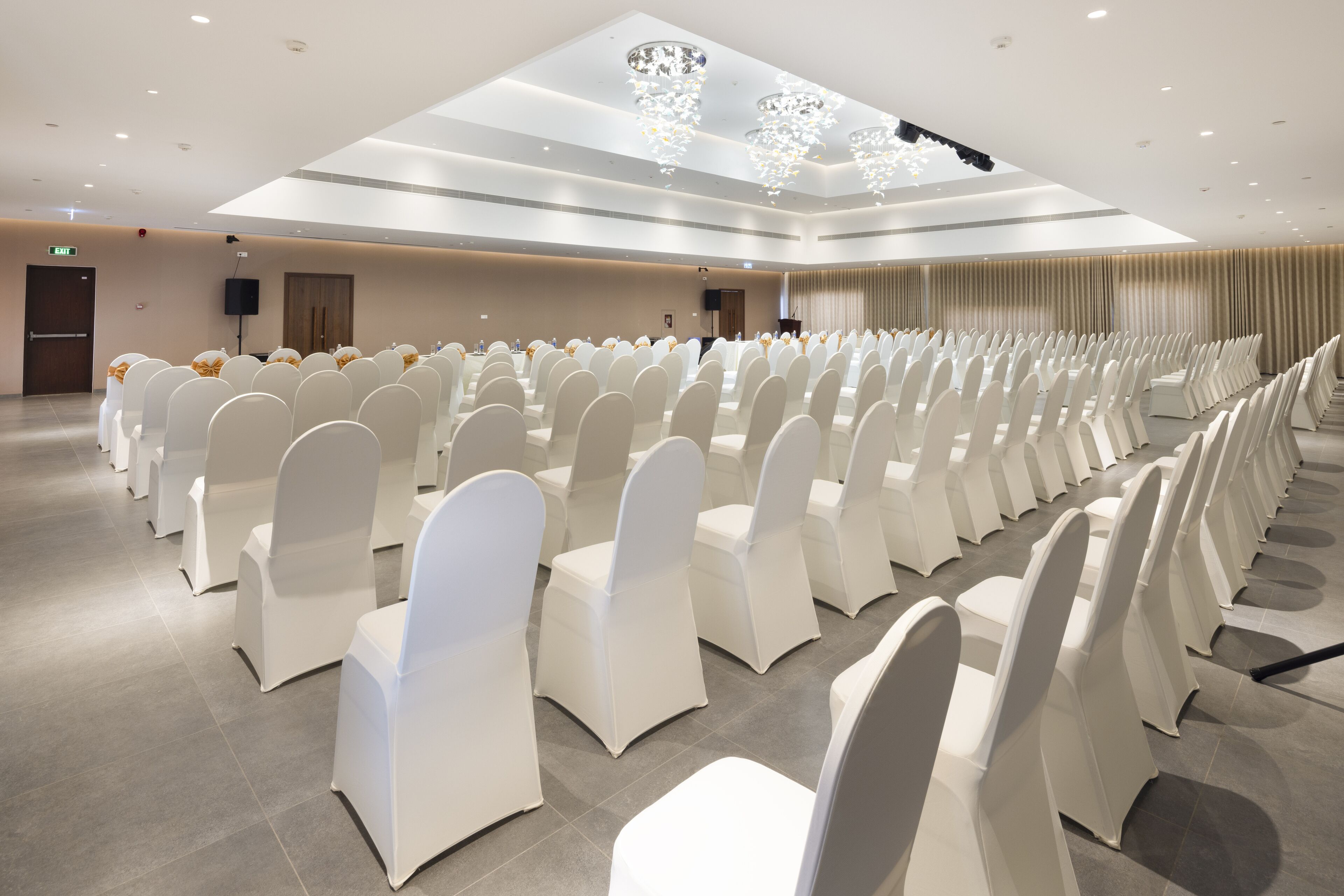 A conference room with rows of white chairs, a stage with a speaker, and a chandelier.