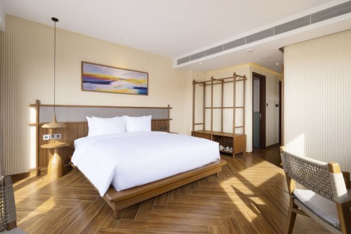 A spacious hotel room with a large bed, wooden flooring, a painting on the wall, and a seating area with a chair and a small table.