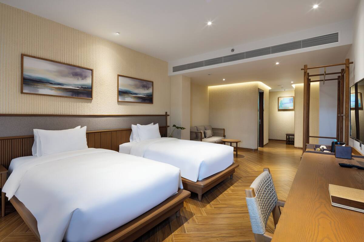 A hotel room with two beds, a wooden desk, a chair, and artwork on the walls.