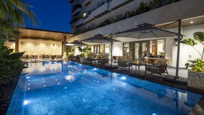 Una piscina con sillones de descanso y sombrillas, rodeada por un edificio con balcones y plantas.