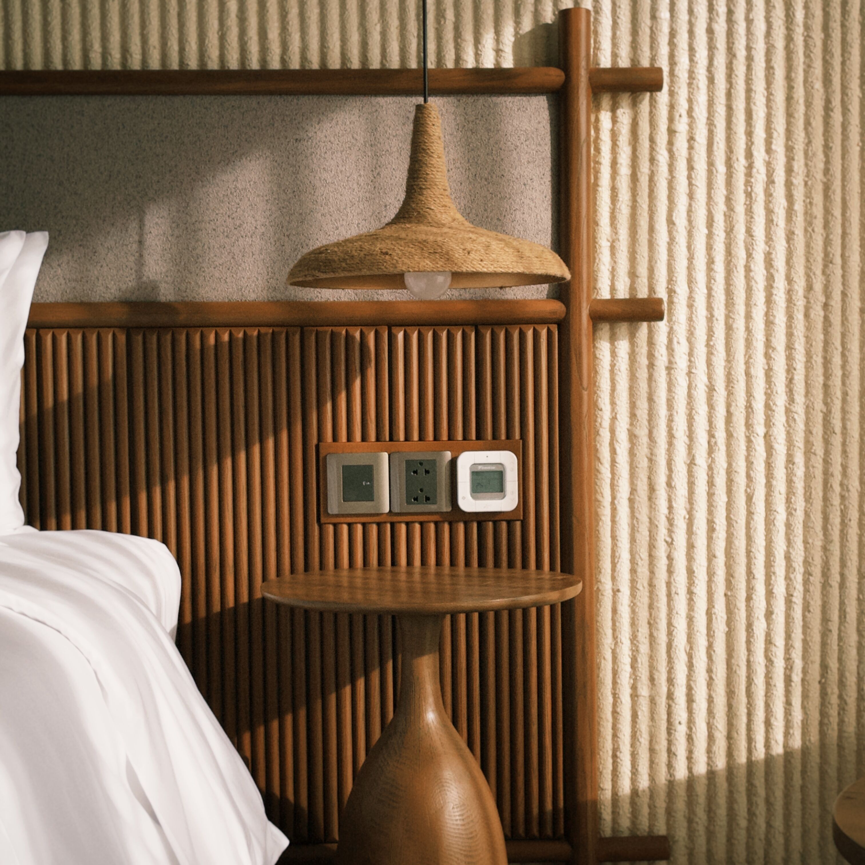 A room with a bed, a small wooden table, a wall-mounted power outlet, and a textured wall.