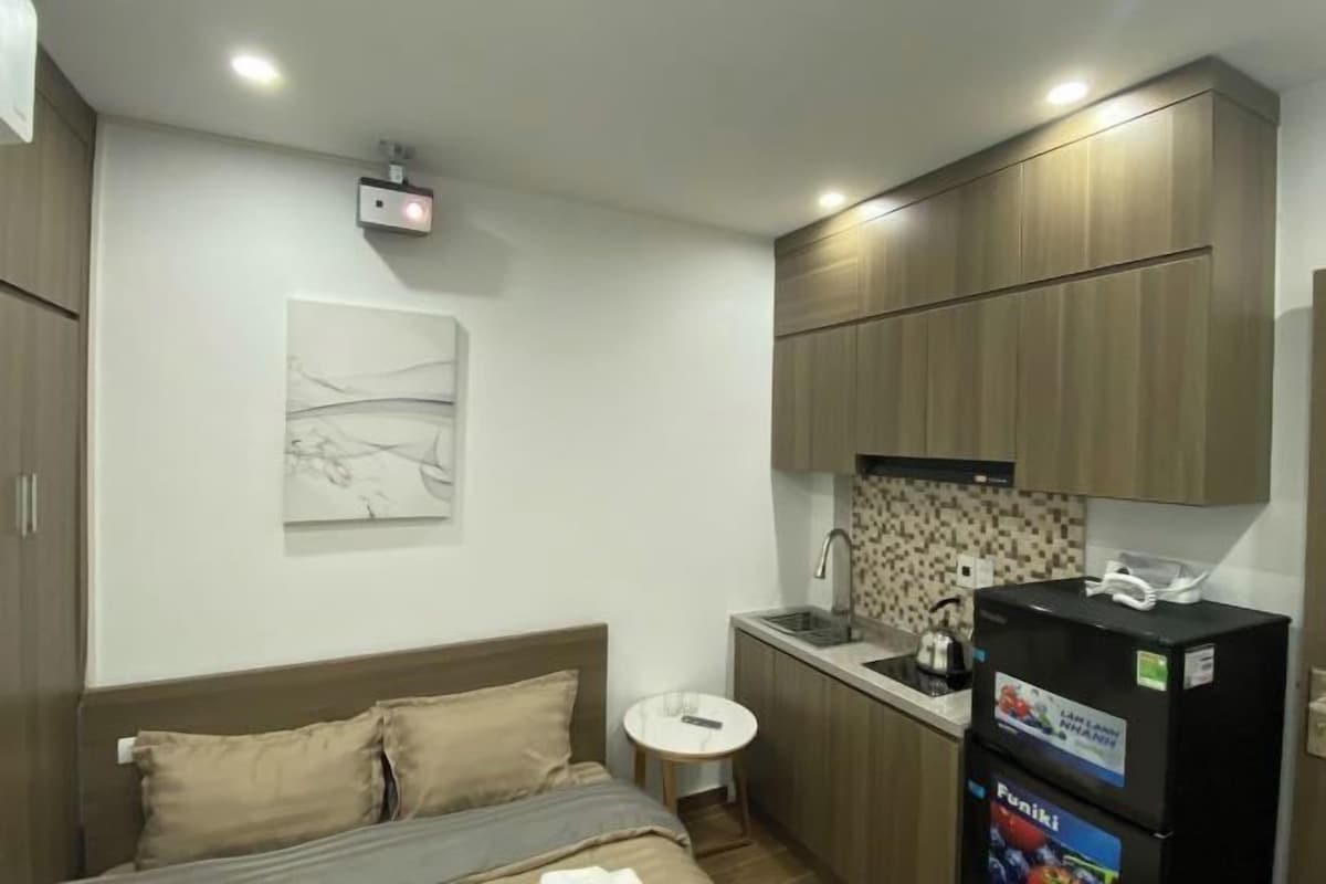A compact hotel room with a bed, a small round table, a kitchenette, and a ceiling-mounted projector.